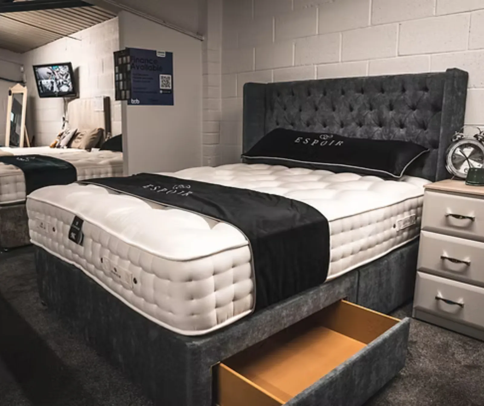 A made up divan bed with a tall buttoned headboard and an open storage drawer on the showroom floor