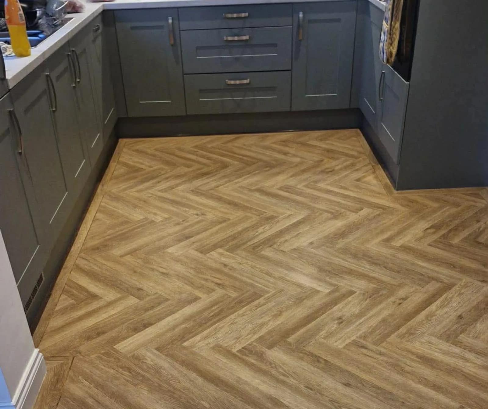 A herringbone wood effect floor laid through a kitchen with grey units and a dark worktop