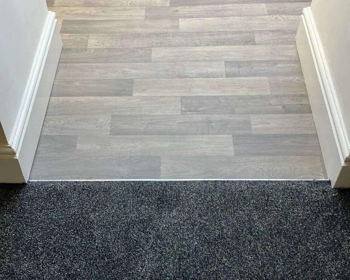 Wood effect flooring meeting a dark carpet at a straight threshold bar
