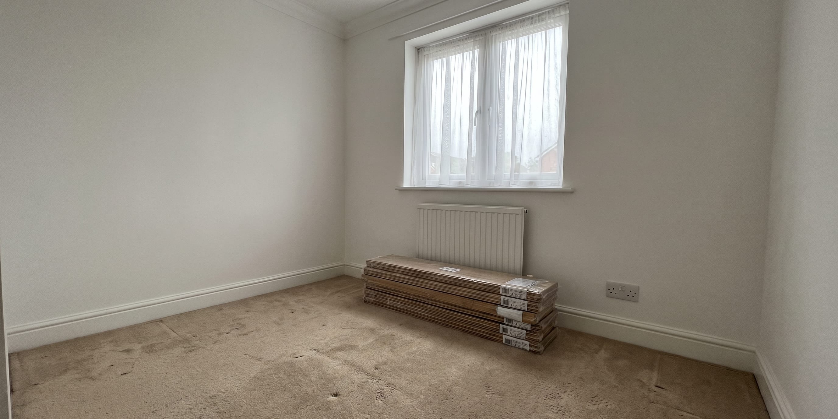 Empty bedroom with beige carpet, a window with net curtains, a wall radiator, and stacked boxes of flooring planks on the floor.
