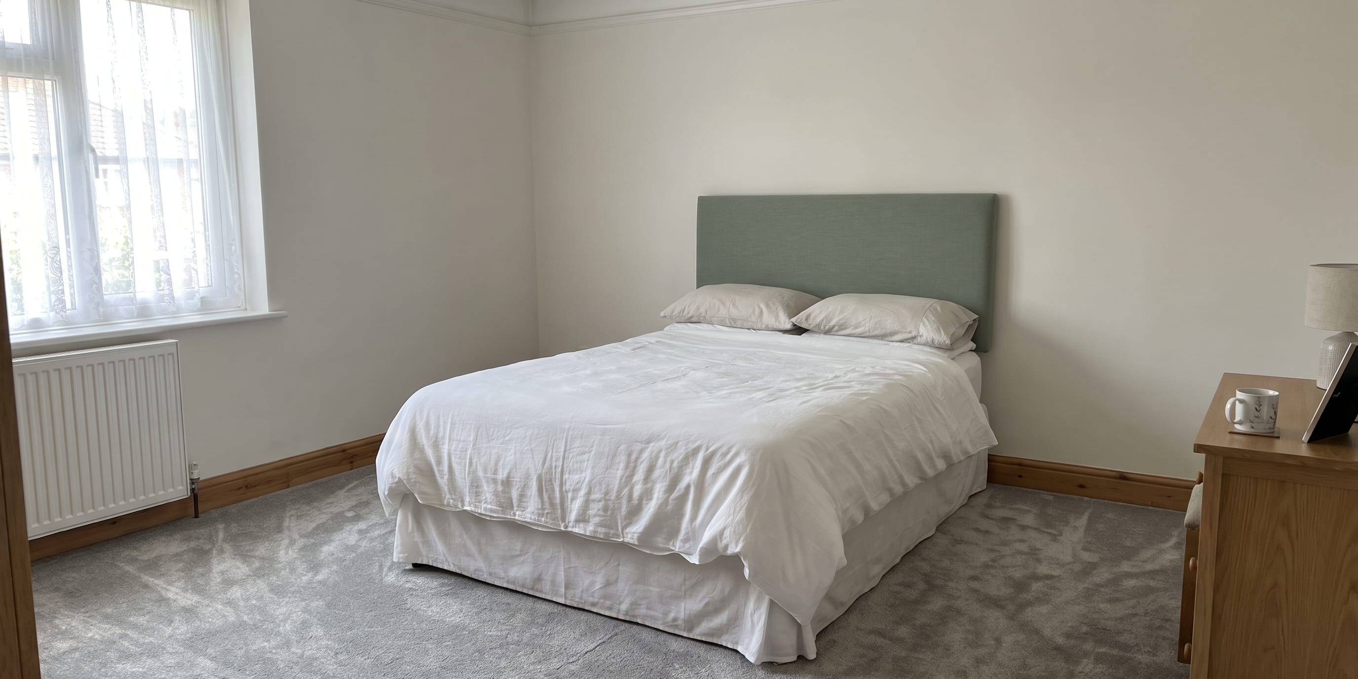 A double bed with white bedding and a sage green upholstered headboard stands against a wall in a carpeted bedroom with a window and wooden dresser.