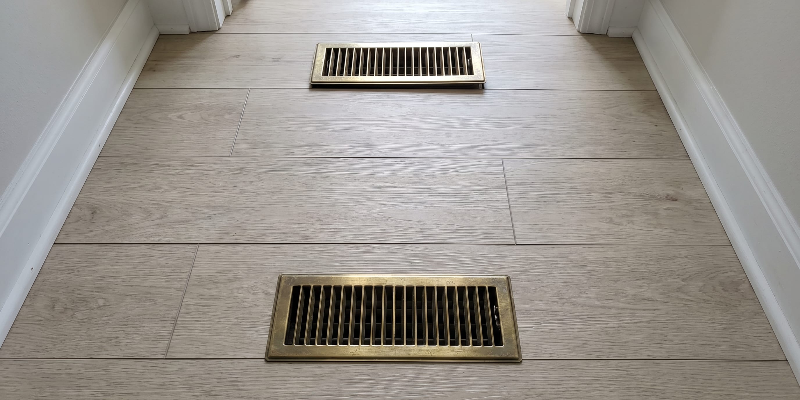 A hallway with wood-look vinyl tile flooring and two brass floor vents, viewed from above.