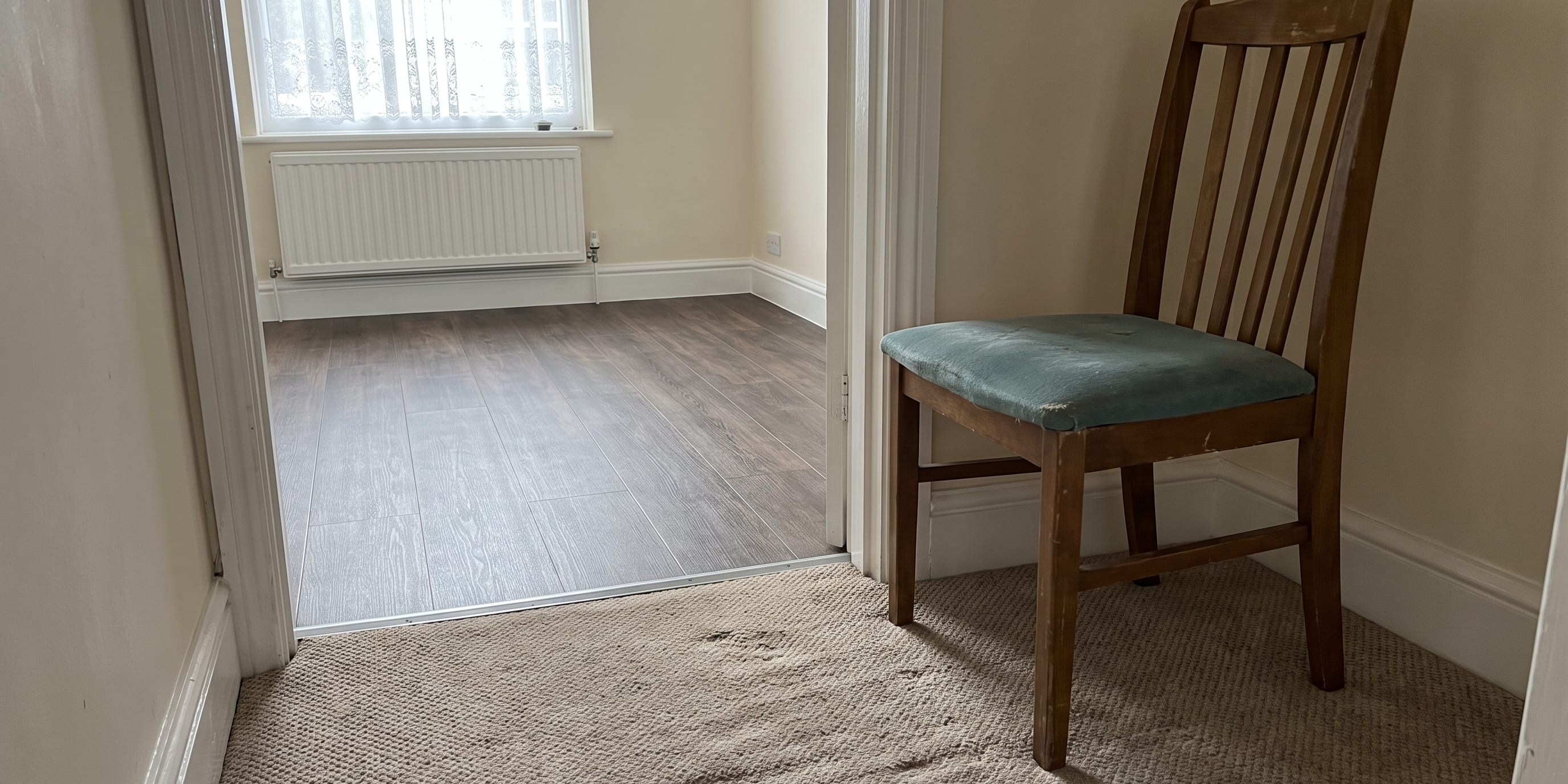 A wooden chair with a worn green cushion sits on beige carpet near a doorway leading to a room with dark laminate flooring and a radiator under a window.