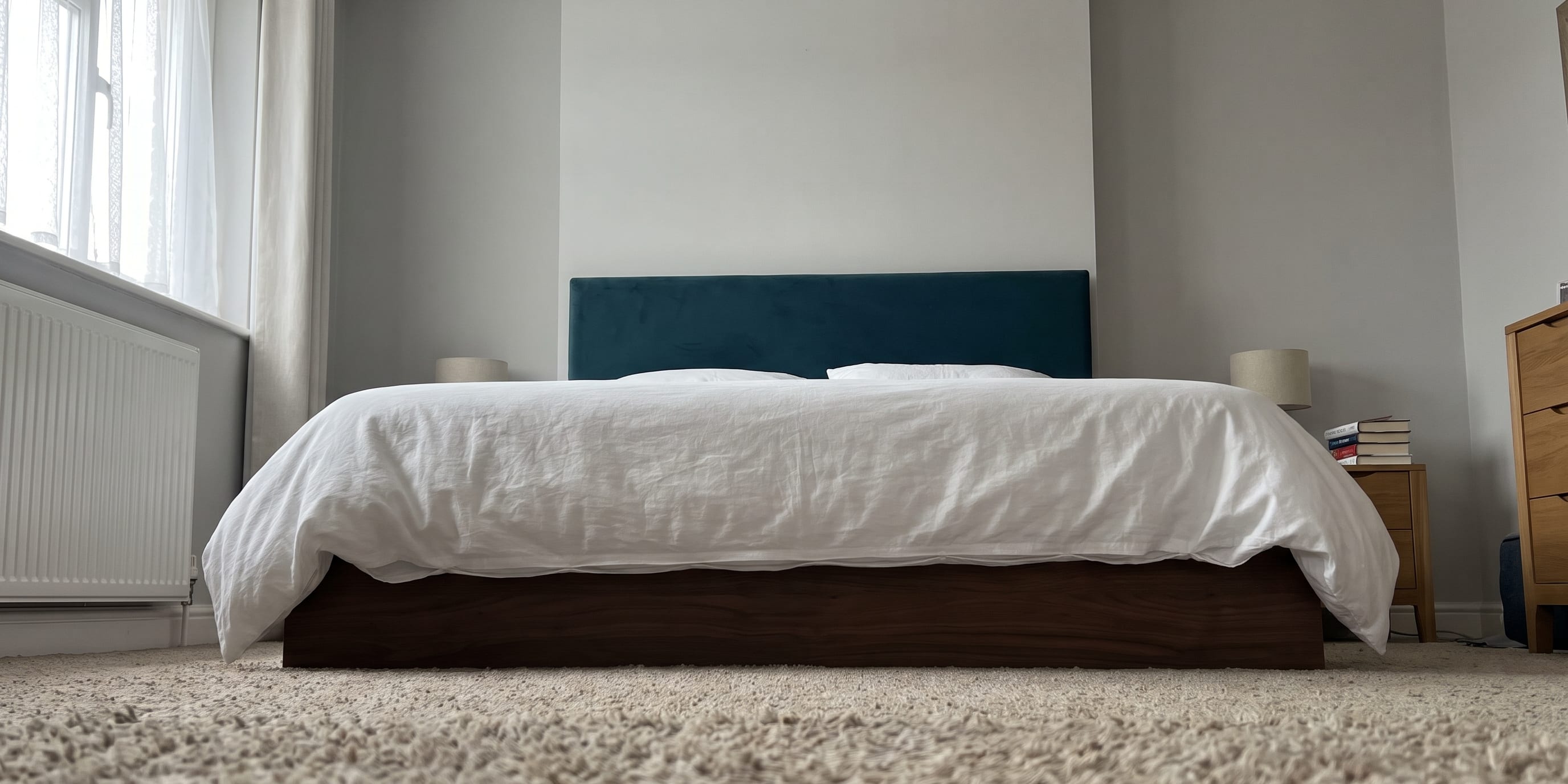 A bed with a teal upholstered headboard and white bedding, photographed low to the carpet, with wooden bedside tables and a radiator visible in a bedroom.