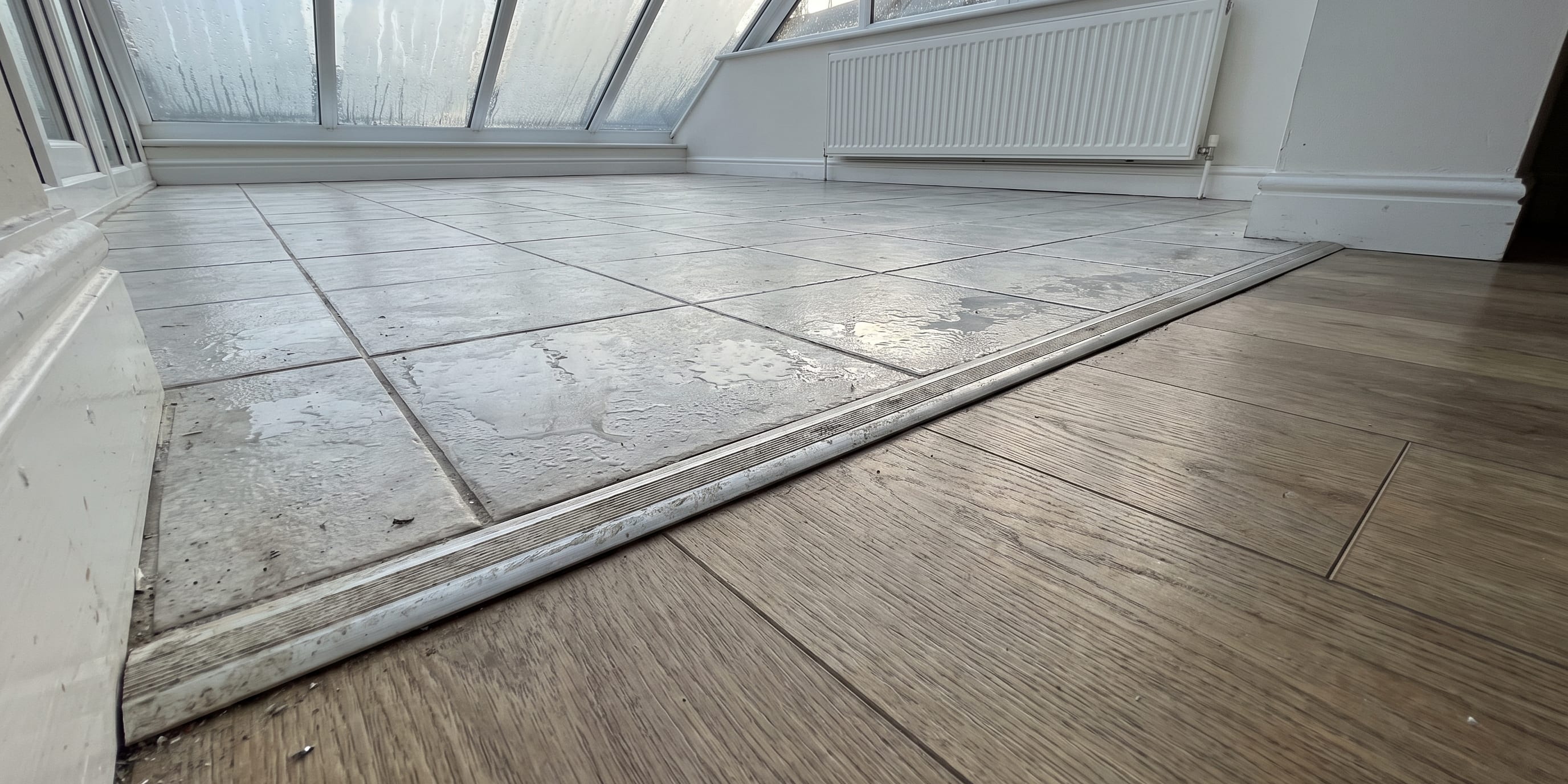 A conservatory floor showing grey tiles transitioning via a metal strip to wood-look flooring, with a radiator under sloped glazed windows.