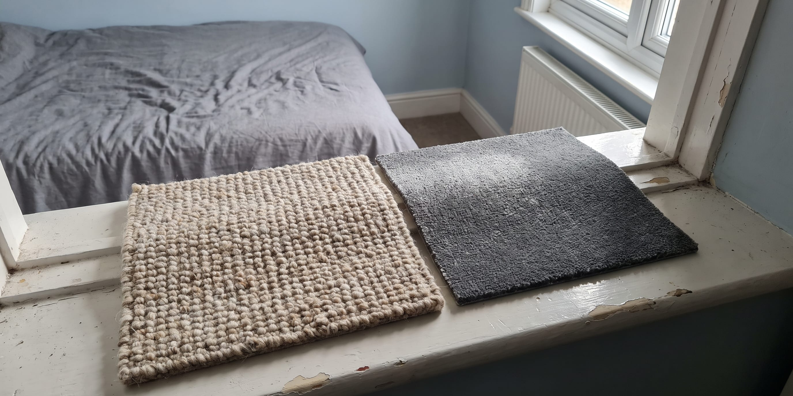 A beige woven wool carpet sample and a dark grey carpet sample lie side by side on a white painted windowsill, with an unmade bed and window visible behind.